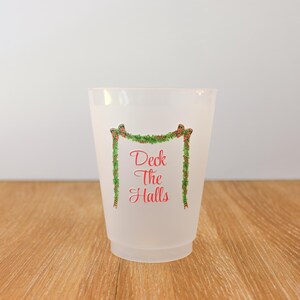 May include: A frosted plastic cup with the text "Deck The Halls" in red script, framed by a green garland with red berries. The cup is on a wooden surface.