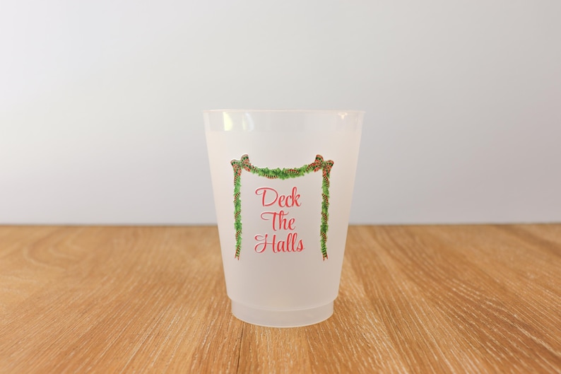 May include: A frosted plastic cup with the text "Deck The Halls" in red script, framed by a green garland with red berries. The cup is on a wooden surface.