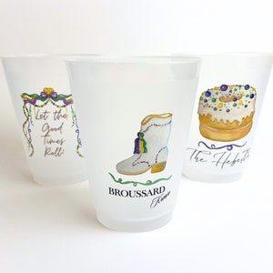 May include: Three clear plastic cups with Mardi Gras themed designs. The first cup has a purple, green, and gold garland with the text "Let the good times roll". The second cup has a white boot with a green tassel and the text "Broussard Krewe". The third cup has a white and gold donut with purple, green, and gold sprinkles and the text "The Hebert's".