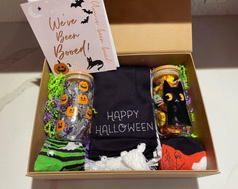 May include: A Halloween gift box with a black scarf that says "Happy Halloween", two glass jars filled with candy, a pair of green and black socks, a pair of red and black socks, and a card that says "We've Been Booed!"