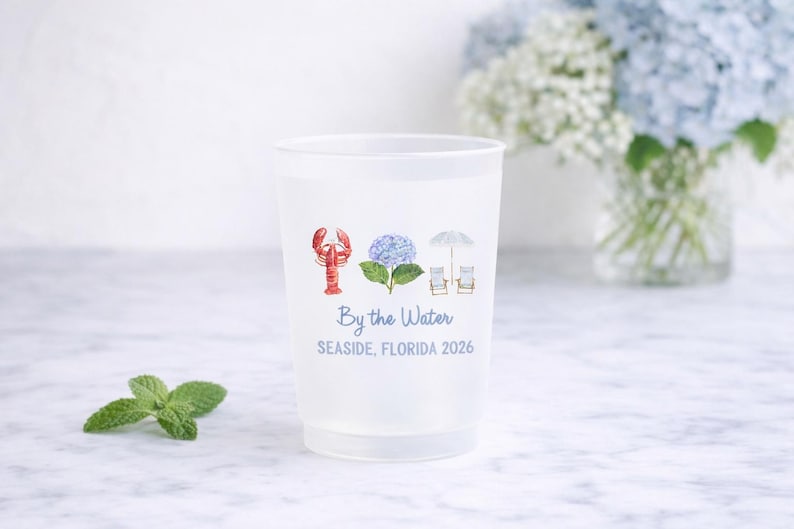 May include: A translucent plastic cup featuring a watercolor-style graphic of a red lobster, a blue hydrangea, and a beach umbrella with two chairs. Below the icons, the text reads By the Water and SEASIDE, FLORIDA 2026 in blue lettering.