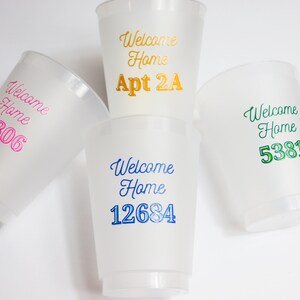 以下が含まれることがあります： 「Welcome Home」の文字と、異なる番号またはマンション番号が印刷された、白いプラスチック製のカップが4つあります。カップは、ピンク、ブルー、グリーン、ゴールドなど、さまざまな色です。