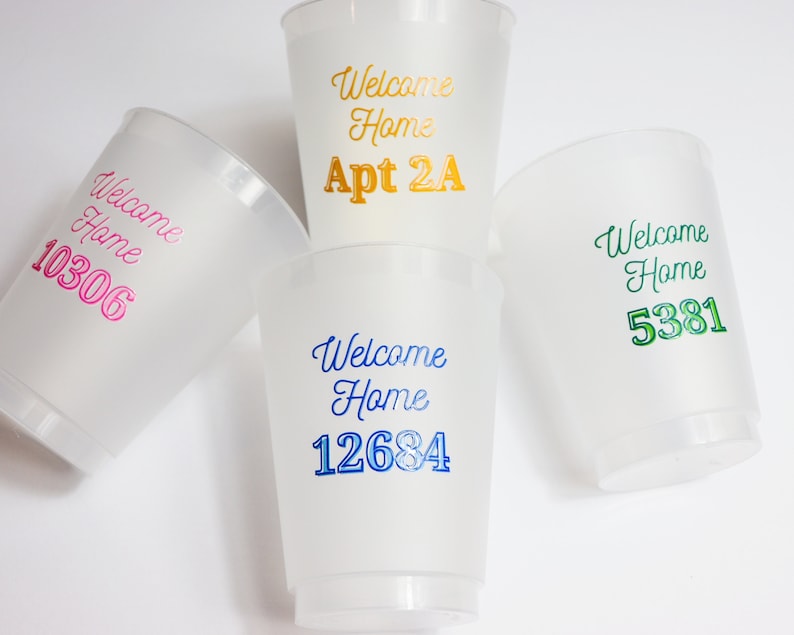 以下が含まれることがあります： 「Welcome Home」の文字と、異なる番号またはマンション番号が印刷された、白いプラスチック製のカップが4つあります。カップは、ピンク、ブルー、グリーン、ゴールドなど、さまざまな色です。
