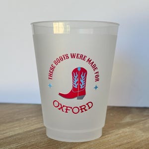 May include: A frosted plastic cup featuring a red cowboy boot graphic with light blue flame details. The text "THESE BOOTS WERE MADE FOR" arches above the boot, while the word "OXFORD" is printed in red below it, flanked by two small blue stars.