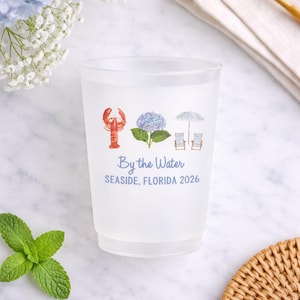 May include: A frosted plastic cup featuring a watercolor graphic of a red lobster, a blue hydrangea, and two beach chairs under an umbrella. The text reads By the Water and SEASIDE, FLORIDA 2026 in blue lettering centered on the front of the cup.
