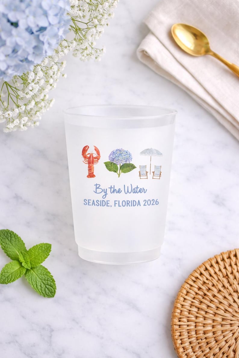 May include: A frosted plastic cup featuring a watercolor graphic of a red lobster, a blue hydrangea, and two beach chairs under an umbrella. The text reads By the Water and SEASIDE, FLORIDA 2026 in blue lettering centered on the front of the cup.