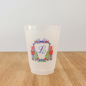 May include: A frosted plastic cup with a festive design. The cup features a shield with the letter "L" in the center, flanked by toy soldiers and Christmas trees. The design is in shades of red, blue, and green.