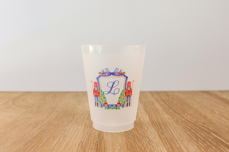 May include: A frosted plastic cup with a festive design. The cup features a shield with the letter "L" in the center, flanked by toy soldiers and Christmas trees. The design is in shades of red, blue, and green.