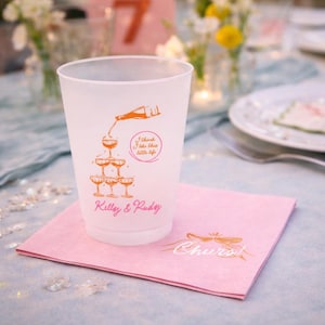 May include: A frosted plastic cup with an orange illustration of champagne being poured into a pyramid of glasses. The cup has the text "I think I like this little life" and the names "Kelly & Rudy". The cup sits on a pink napkin.