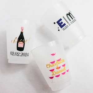 May include: Three white plastic cups with gold and pink designs. The first cup has a champagne bottle with the text "cheers!" and "02.02.2024". The second cup has a stack of pink champagne glasses with the text "Charlotte & Daniel". The third cup has the text "E|M Eliza & Richard".