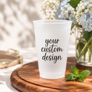 May include: A white, frosted plastic cup with the text "your custom design" in black script. The cup sits on a wooden serving board with a sprig of mint. A vase of blue and white flowers is in the background.
