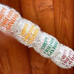 May include: Four clear glass tumblers with the text "Happy Friends Giving" in orange, yellow, green, and red lettering. The tumblers are stacked on top of each other and surrounded by white packing paper.