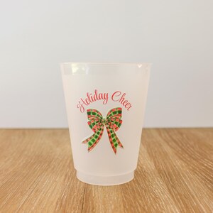 May include: A frosted plastic cup with the words "Holiday Cheer" in red script above a watercolor bow. The bow has red, green, and gold plaid details. The cup is on a wooden surface.