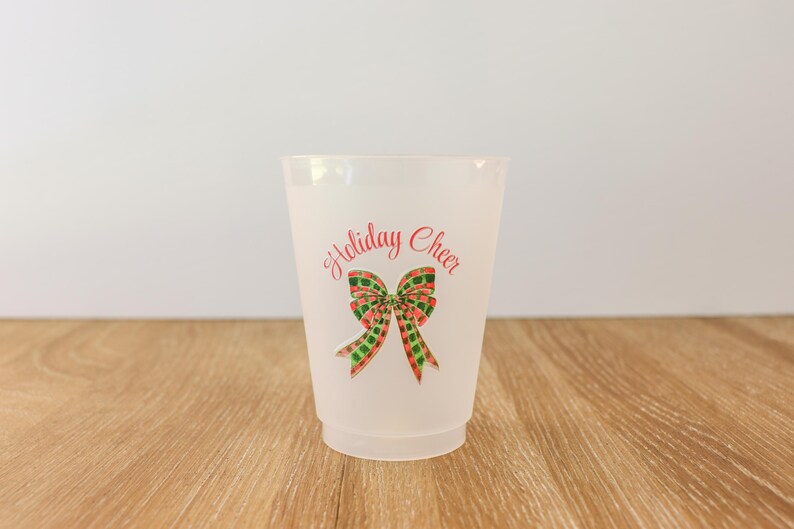 May include: A frosted plastic cup with the words "Holiday Cheer" in red script above a watercolor bow. The bow has red, green, and gold plaid details. The cup is on a wooden surface.
