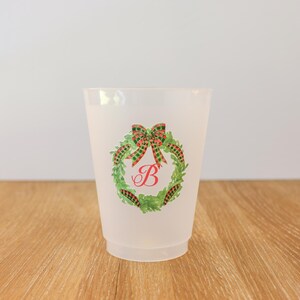 May include: A frosted plastic cup featuring a Christmas wreath design. The wreath is green with a red and green plaid bow and a red letter "B" in the center. The cup is sitting on a wooden surface.