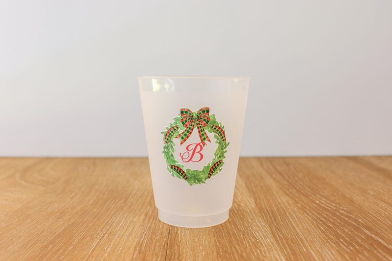 May include: A frosted plastic cup featuring a Christmas wreath design. The wreath is green with a red and green plaid bow and a red letter "B" in the center. The cup is sitting on a wooden surface.