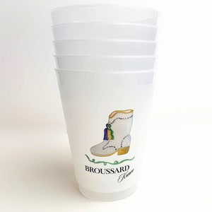 May include: Stack of five white plastic cups with a design of a white boot with gold and purple accents. The boot has a green tassel and the text "BROUSSARD Krewe" is printed below the boot.