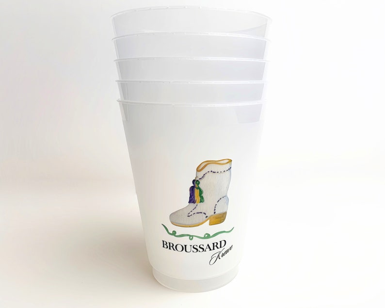 May include: Stack of five white plastic cups with a design of a white boot with gold and purple accents. The boot has a green tassel and the text "BROUSSARD Krewe" is printed below the boot.