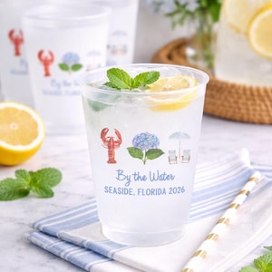 May include: A clear plastic cup filled with a beverage, a lemon slice, and mint leaves. The cup features a printed design of a red lobster, a blue hydrangea, and two beach chairs under an umbrella, with the text By the Water SEASIDE, FLORIDA 2026.