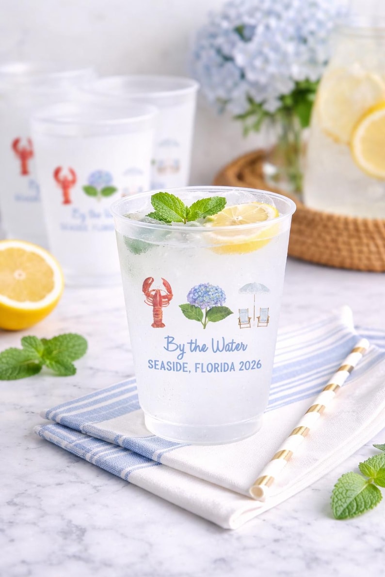 May include: A clear plastic cup filled with a beverage, a lemon slice, and mint leaves. The cup features a printed design of a red lobster, a blue hydrangea, and two beach chairs under an umbrella, with the text By the Water SEASIDE, FLORIDA 2026.
