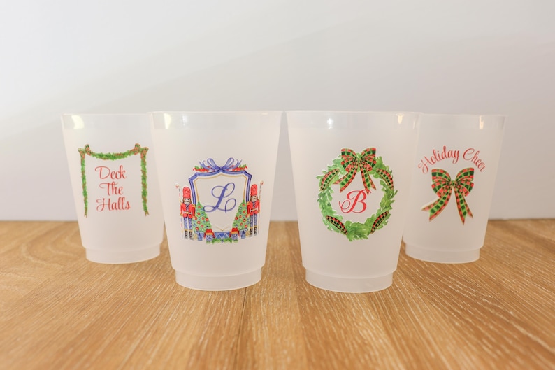 May include: Four frosted plastic cups with holiday-themed designs. One cup has "Deck the Halls" text, another features a crest with the letter "L", a third has a wreath with the letter "B", and the last says "Holiday Cheer" with a bow.