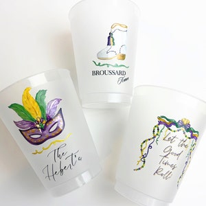 May include: Three clear plastic cups with Mardi Gras themed designs. The first cup has a purple, gold, and green mask with feathers. The second cup has a white boot with gold trim and the text "Broussard Family". The third cup has a purple, gold, and green garland with the text "Let the good times roll!"