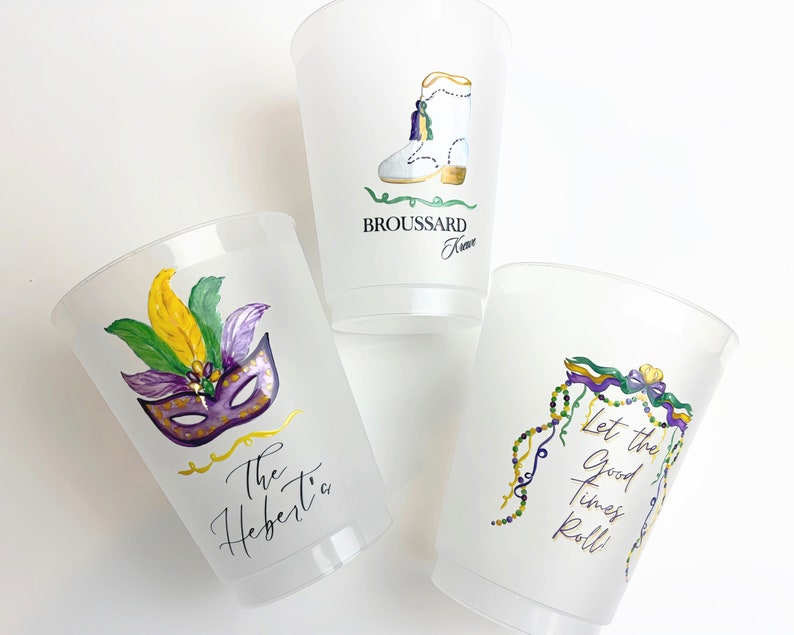 May include: Three clear plastic cups with Mardi Gras themed designs. The first cup has a purple, gold, and green mask with feathers. The second cup has a white boot with gold trim and the text "Broussard Family". The third cup has a purple, gold, and green garland with the text "Let the good times roll!"