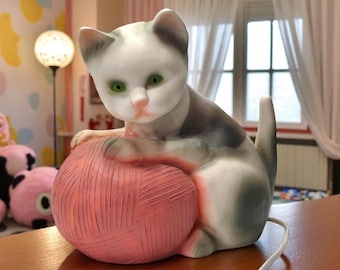 May include: A ceramic cat ornament with green eyes and a pink nose, sitting on a pink ball of wool. The cat has grey and white markings. A white cord extends from the base. The lamp is on a wooden surface.