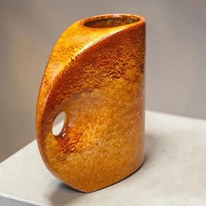 Può includere: Un vaso in ceramica con una smaltatura maculata color ambra e marrone. L'oggetto presenta un profilo laterale curvo distintivo con un'impugnatura circolare ritagliata integrata nel corpo, che porta a un'apertura stretta di forma ovale.