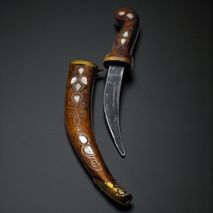 May include: An antique dagger with a curved blade and wooden handle. The handle and sheath are adorned with inlaid mother-of-pearl and gold accents. The dagger is displayed against a dark background, showcasing its intricate design and craftsmanship.