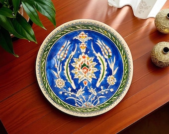 Puede incluir: Un plato decorativo de cerámica con un vibrante diseño floral. El plato presenta un centro azul profundo con motivos florales blancos, verdes, amarillos y rojos. Un borde verde rodea el diseño central, y el borde es blanco con un patrón repetido.