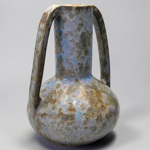 May include: A ceramic vase with a bulbous base and a tall, narrow neck featuring two large, arched handles. The surface has a mottled glaze finish in shades of light blue, grey, and earthy brown, creating a textured, variegated appearance.