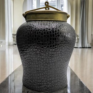 Op de afbeelding: Een zwarte keramische pot met een textuur die lijkt op krokodillenleer. De pot heeft een gouden deksel met een decoratief handvat.