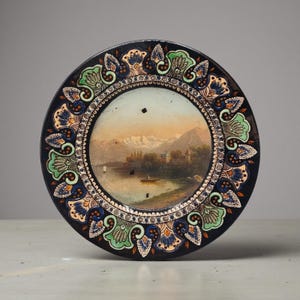 Può includere: Un piatto decorativo rotondo in ceramica con un paesaggio centrale dipinto raffigurante un lago, una piccola barca e montagne innevate in lontananza. L'ampio bordo è decorato con un motivo ripetitivo di fiori verdi, blu e arancioni su nero.