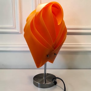 Op de afbeelding: Een tafellamp met een feloranje kap opgebouwd uit overlappende, gebogen kunststof vellen die zijn vastgezet met twee kleine zilverkleurige metalen klinknagels. De kap wordt ondersteund door een slanke, verchroomde metalen stang en voet.