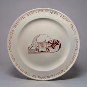 Puede incluir: Un plato de cerámica blanco con una ilustración marrón de dos rostros estilizados y líneas abstractas. El borde del plato presenta el texto "canova. soieries de Lyon. andré claude" en un patrón repetitivo.