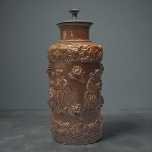 May include: A brown salt-glazed stoneware tankard featuring a cylindrical body with relief-moulded figures of people in historical attire, floral motifs, and a decorative border. The vessel includes a matching fitted lid with a central knob.