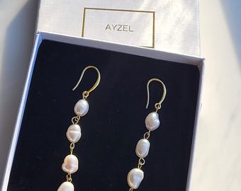 Può includere: Un paio di orecchini dorati con gocce di perle bianche. Gli orecchini sono in una scatola bianca con la scritta "AYZEL" stampata sul coperchio.