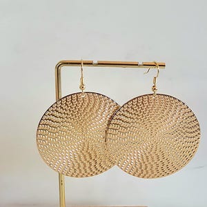 Può includere: Un paio di orecchini circolari color oro con una superficie strutturata e fantasia. Gli orecchini sono esposti su un supporto per orecchini color oro su sfondo bianco. Gli orecchini hanno una chiusura a gancio.
