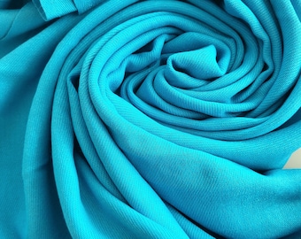 Könnte beinhalten: Nahaufnahme eines leuchtend türkisfarbenen Stoffes, kunstvoll in wirbelnden Falten angeordnet. Der Stoff hat eine subtile Textur mit sichtbaren Webdetails. Die leuchtend blaue Farbe dominiert den Rahmen und erzeugt ein Gefühl von Tiefe und Bewegung. Der Stoff wirkt weich und fließend.