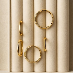 May include: Five gold wedding bands are displayed on a beige, vertically-ribbed surface. The rings are positioned in various orientations, showcasing their circular design and polished finish. The lighting highlights the rings' metallic lustre.