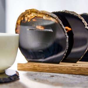 May include: A set of four black agate coasters with gold edges, displayed on a wooden holder. A white teacup sits to the left, with a coaster underneath. The coasters have a natural, organic shape and are a decorative home accessory.