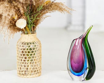 May include: A glass vase with a woven rattan sleeve contains dried flowers and grasses. Next to it, a colourful art glass vase displays layers of purple, blue, and green. A decorative home decor item.
