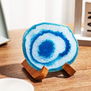 May include: A blue and white agate slice displayed on a small wooden stand. The agate features concentric rings of color, with a deep blue center and a light blue and white outer edge. The wooden stand has a natural finish.