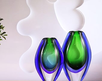 May include: Two decorative glass vases. The vases have a clear base, a vibrant green interior, and are outlined with a deep blue. The vases are different sizes and sit on a wooden surface. The background features a white, abstract design.