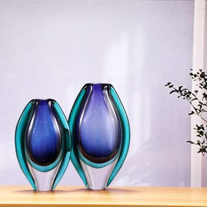 May include: Two glass vases with a teardrop shape. The vases have a gradient of colors, with a clear base, a teal outer layer, and a deep blue interior. They are displayed on a wooden surface against a white background.