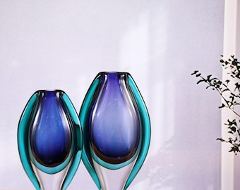 May include: Two glass vases in a teardrop shape. The vases feature a colour gradient, with a clear base, a teal outer layer, and a deep blue interior. They are displayed on a wooden surface against a white background.