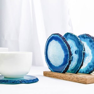 May include: A white teacup sits on a blue and white agate coaster. Three additional agate coasters in a wooden holder are visible. The coasters have a blue and white color scheme, with a natural, organic edge.