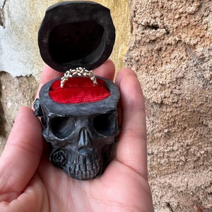Puede incluir: Una caja de anillo de calavera negra con un interior de terciopelo rojo. Un anillo de plata se muestra dentro de la caja.