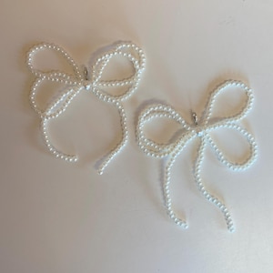 May include: Two white pearl bow tie hair clips. Each clip is made of a string of white pearls that are tied into a bow shape.